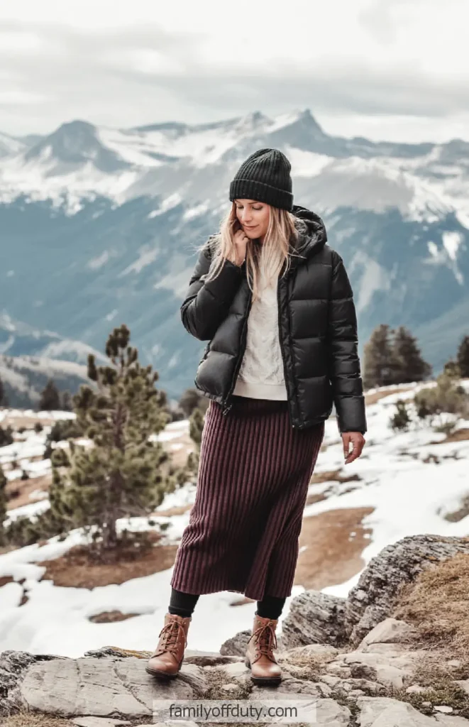 Woman in winter attire standing on snowy mountain path with a scenic view in the background.