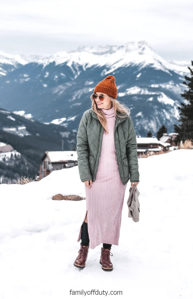 Woman in winter clothing poses with mountains in background, wearing a green jacket, pink dress, and orange beanie.