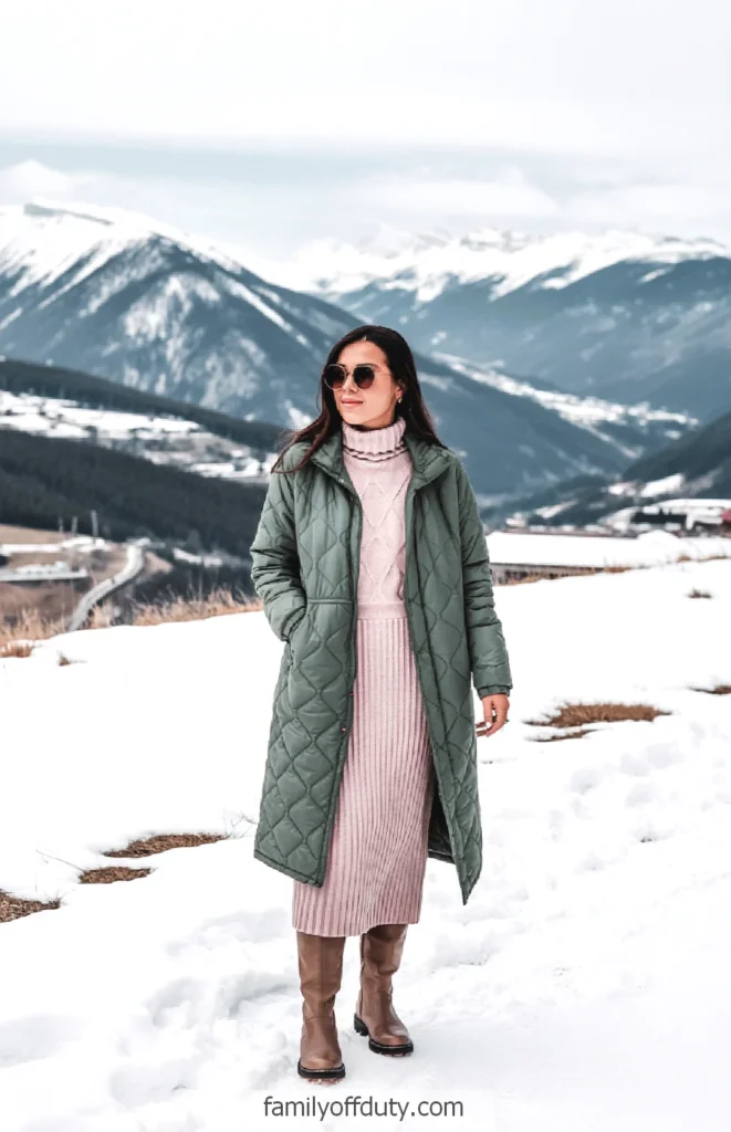 Woman in green winter coat and pink dress stands in snowy mountain landscape.