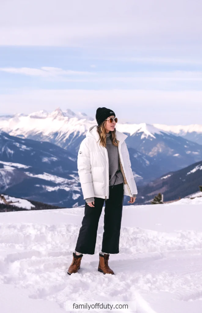 Person in winter outfit enjoys snowy mountain view. Wearing a white jacket, beanie, and sunglasses.