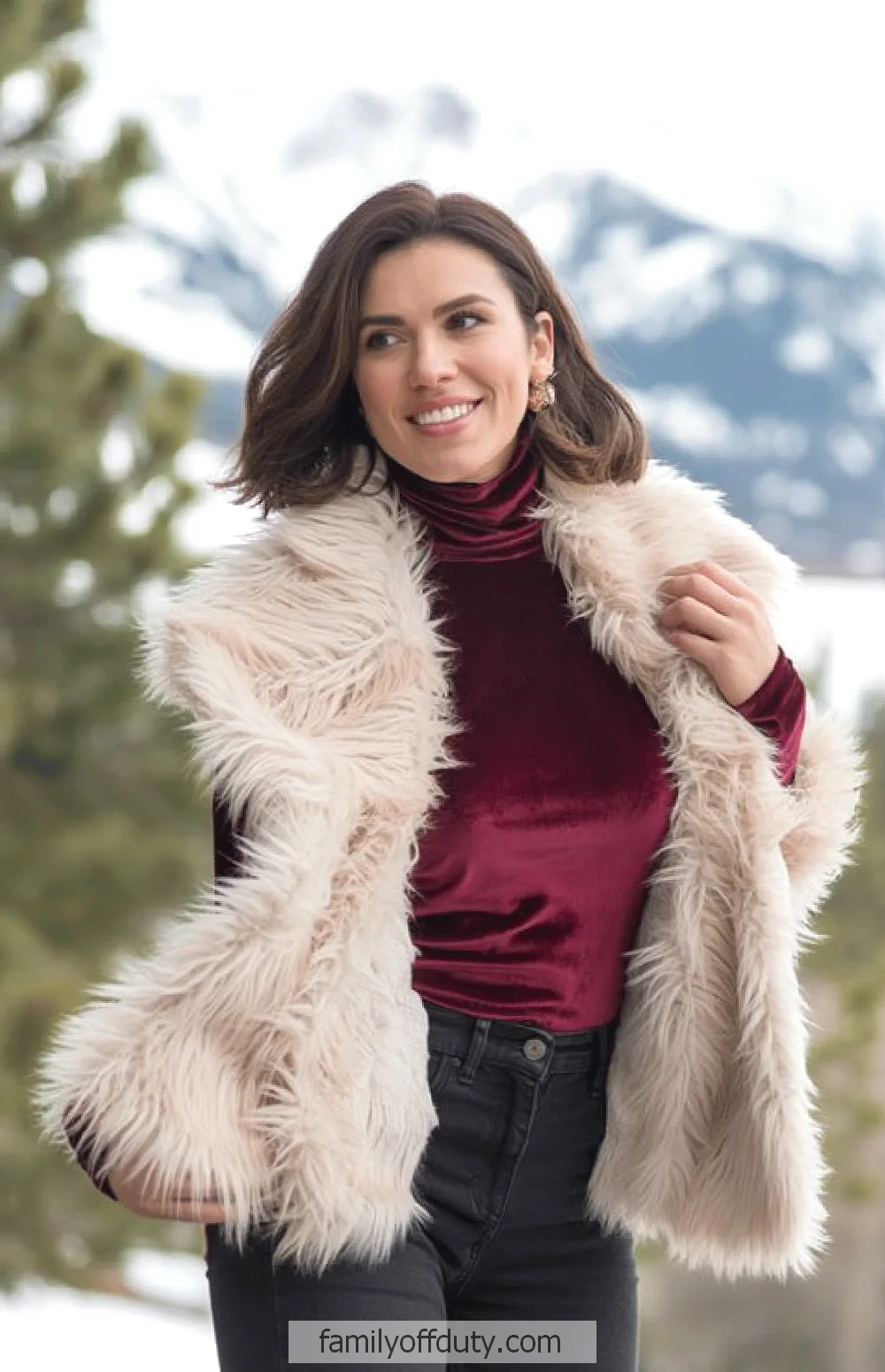Mountain winter outfit ideas. Woman in faux fur vest and red top smiles outdoors with snowy mountain background. Fashion and winter style.