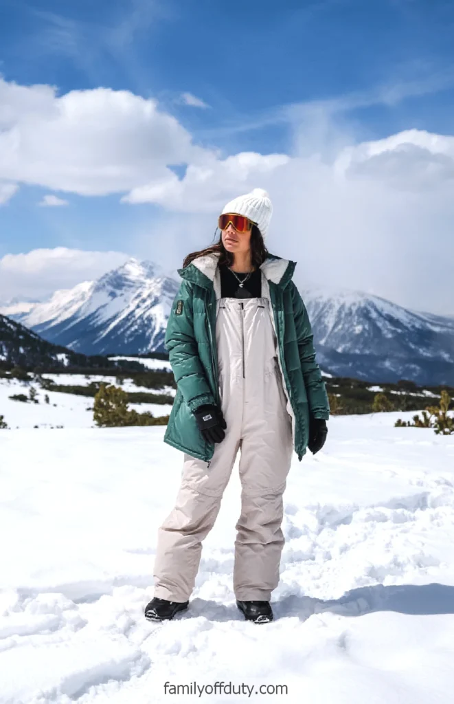 Person in winter gear stands in snowy landscape with mountains in the background.