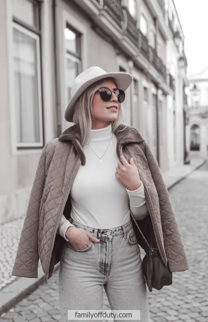Stylish woman in sunglasses and hat wearing a quilted jacket, white top, and jeans on a city street.