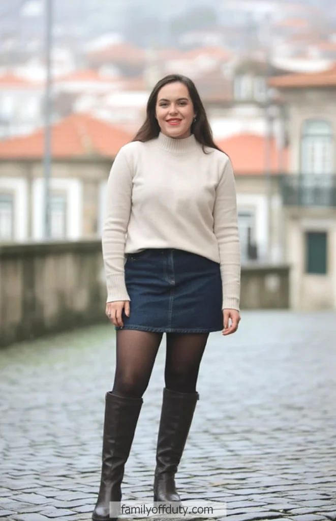 Person wearing a beige sweater, denim skirt, and boots on a cobblestone street in a quaint town setting.