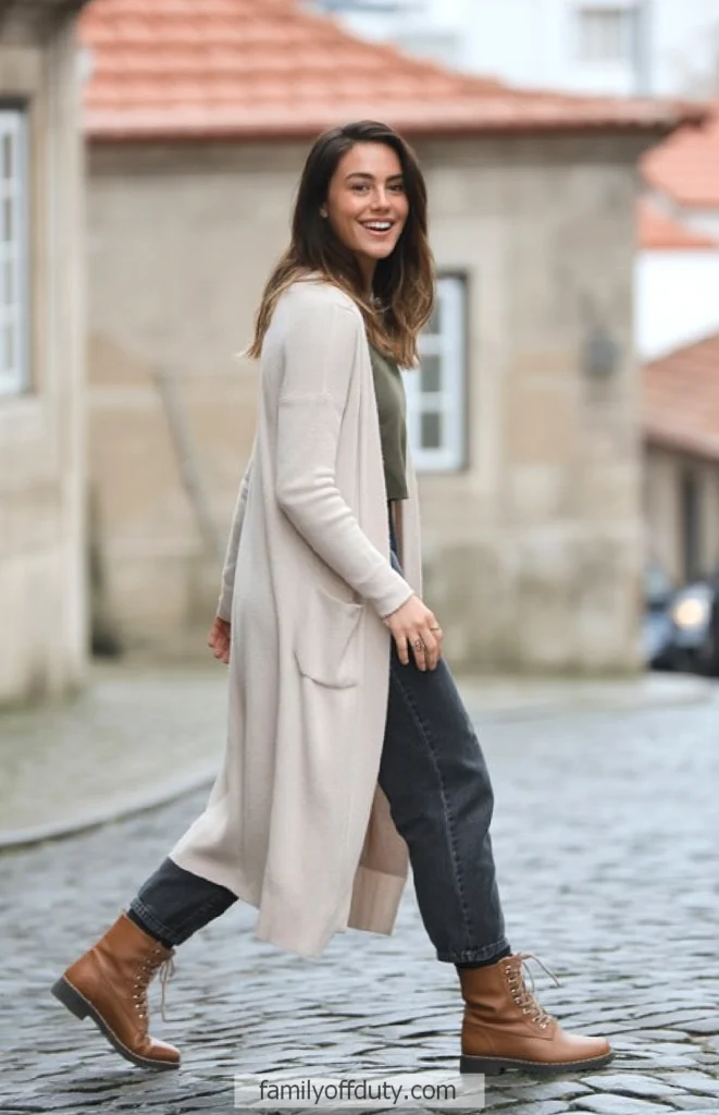 Portugal winter outfit ideas. Woman in casual outfit walking on cobblestone street, smiling, wearing a long beige cardigan and brown boots.