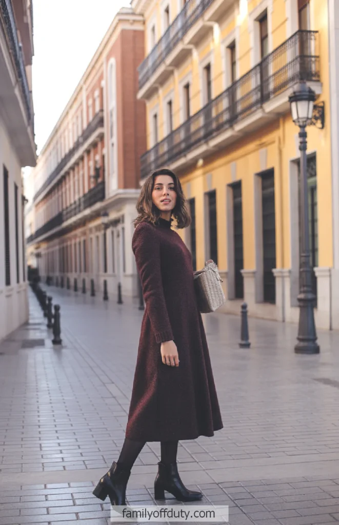 Woman in a burgundy dress and boots walking down a European-style city street with colorful buildings.