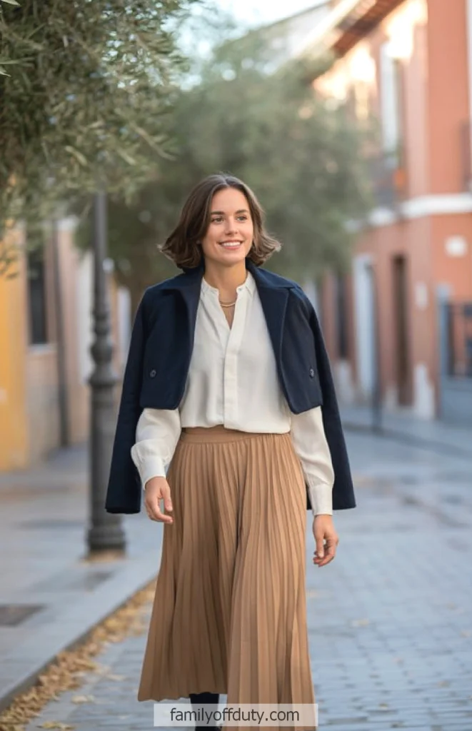 Smiling woman walking in stylish fall outfit, pleated skirt and navy coat, on a city street.