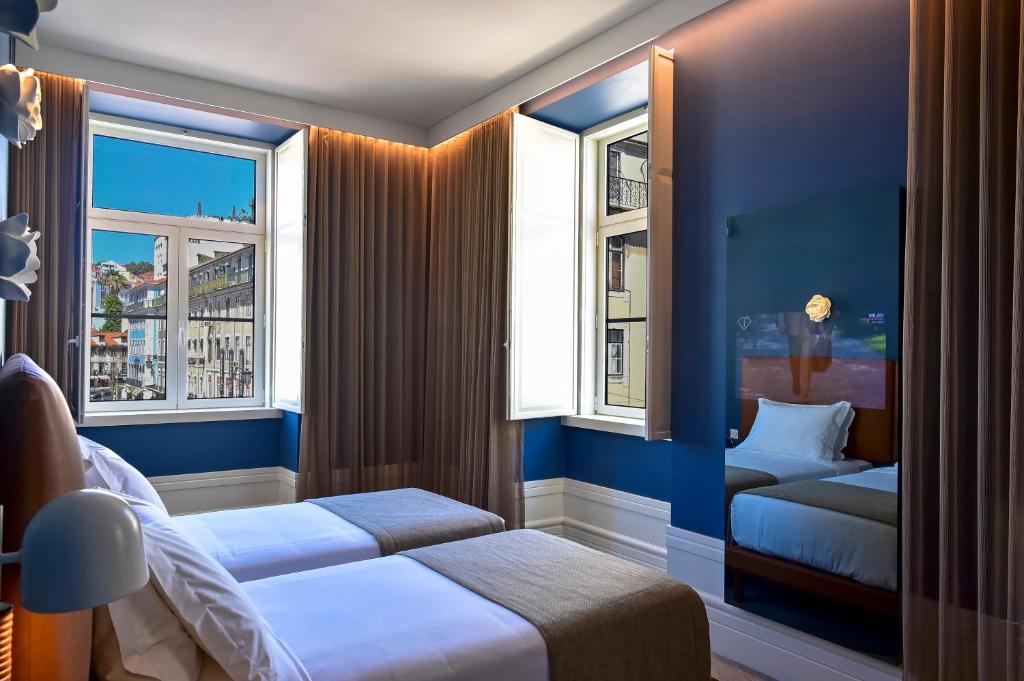 Modern hotel room with two beds, blue walls, and city view through large windows. Cozy and inviting atmosphere.