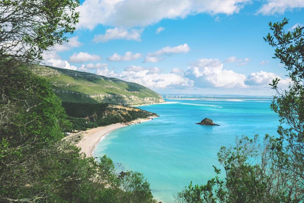 Scenic coastal view with blue sea, sandy beach, and lush green cliffs under a bright sky with fluffy clouds.