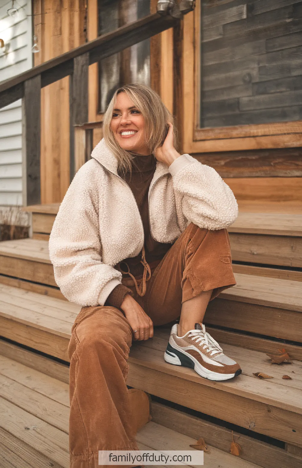cabin fall outfit ideas. Cozy autumn outfit with a woman smiling in a sherpa jacket and sneakers, sitting on wooden steps.