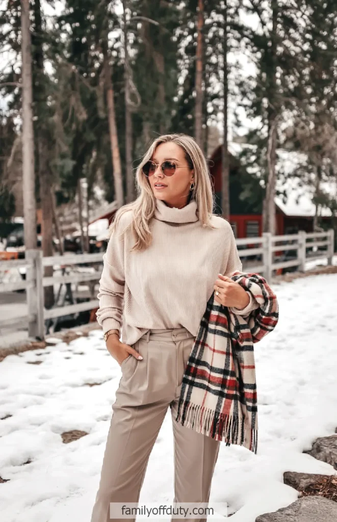 Casual winter fashion: Woman in beige turtleneck, plaid scarf, beige pants in snowy outdoor setting.