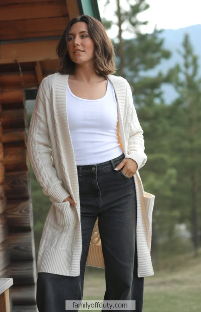 Woman in white cardigan and jeans standing outside a cabin in a forest setting.