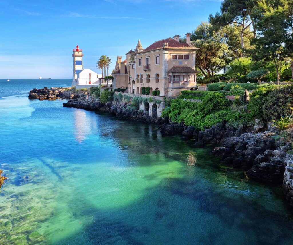 Scenic coastal view with a historic building and lighthouse by the sea, surrounded by clear blue waters and lush greenery.