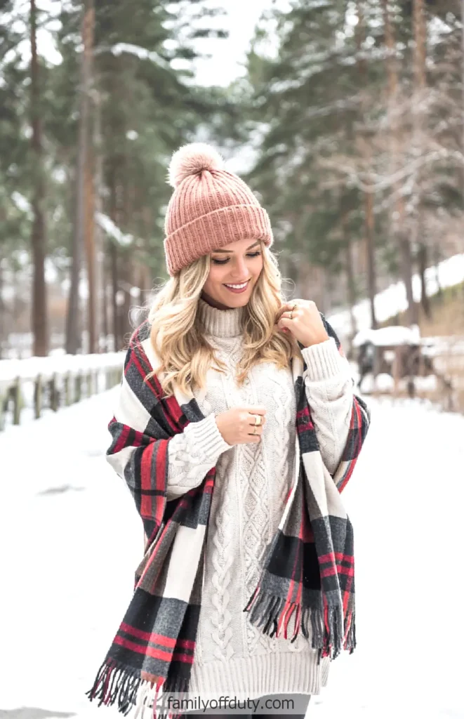 Woman in cozy winter attire with pink beanie, plaid scarf in snowy forest setting.