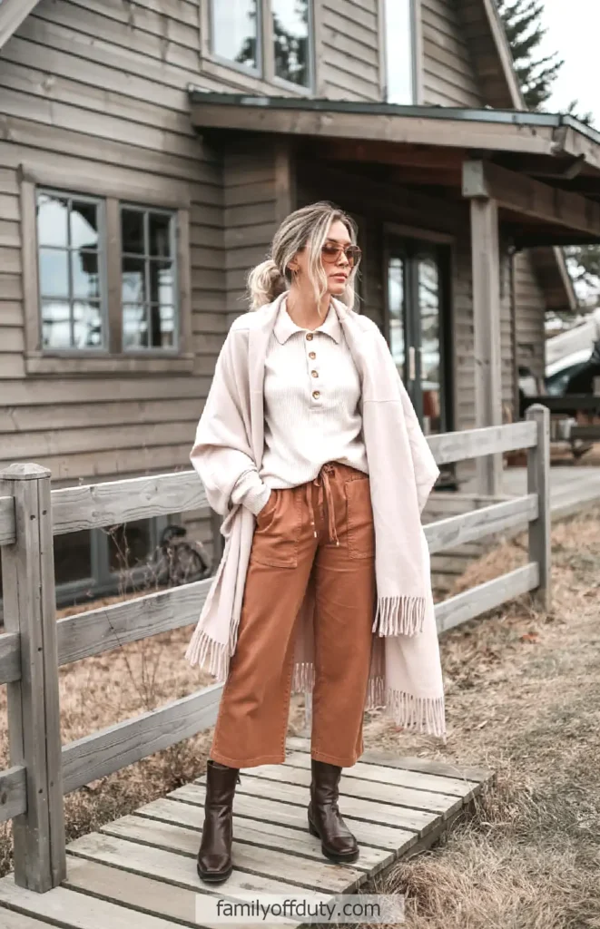Woman in cozy fall outfit with sunglasses and scarf, standing on wooden path by rustic cabin.
