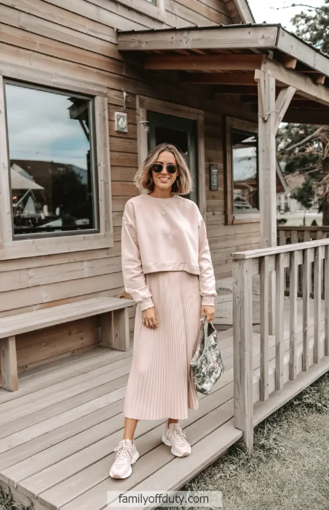 Woman in pink outfit smiling in front of a rustic wooden cabin, holding a floral bag.
