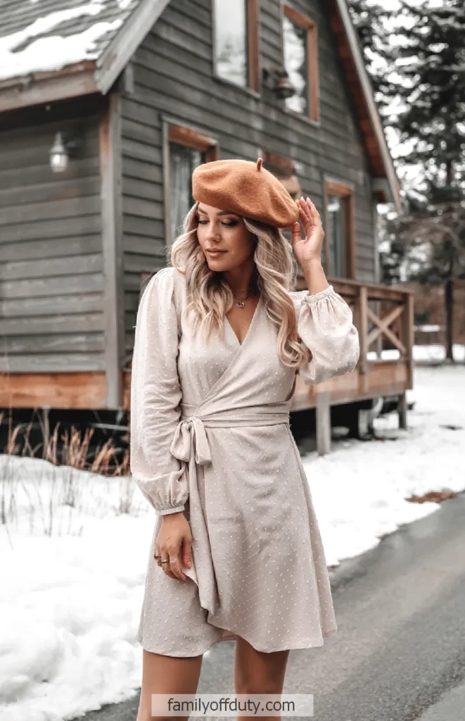 Woman in beige dress and beret poses outdoors by a snowy cabin.