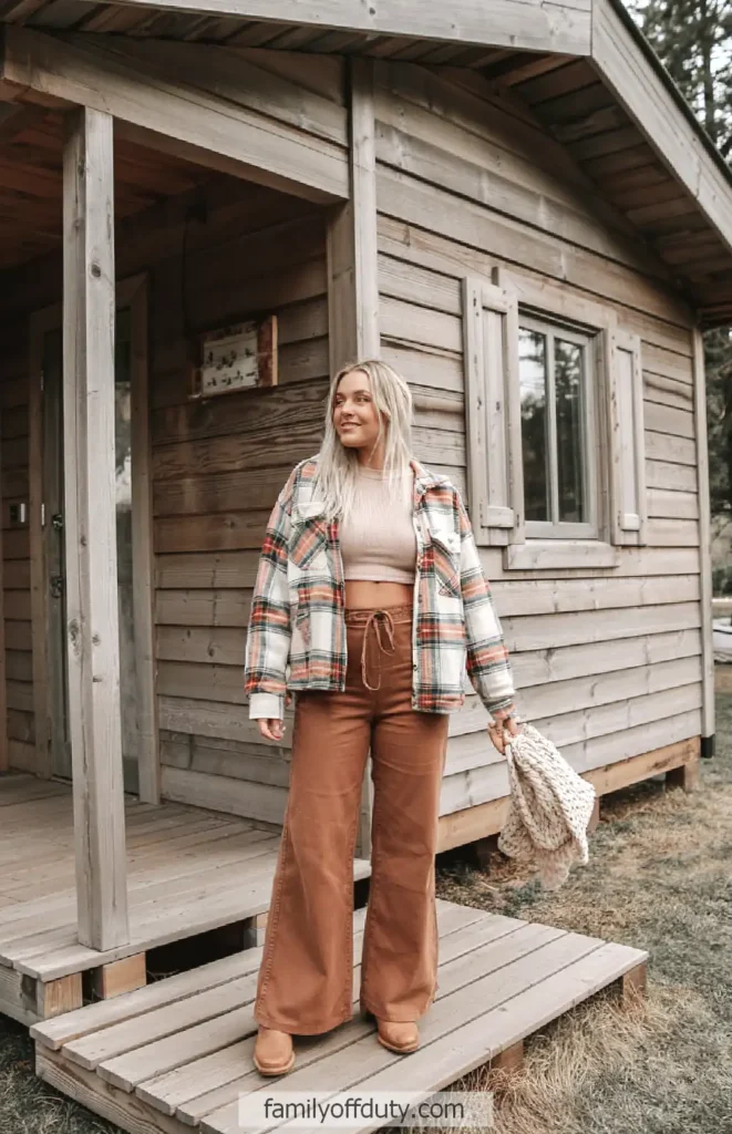 Woman in plaid jacket and brown pants standing outside a wooden cabin.