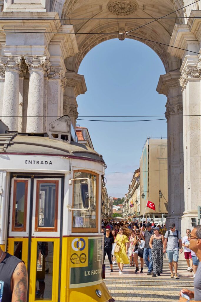 Yellow tram passing under Lisbon's Rua Augusta Arch, bustling with tourists on a sunny day.