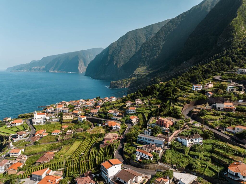 Aerial view of coastal village with red-roofed houses, lush greenery, and dramatic cliffs by the ocean.