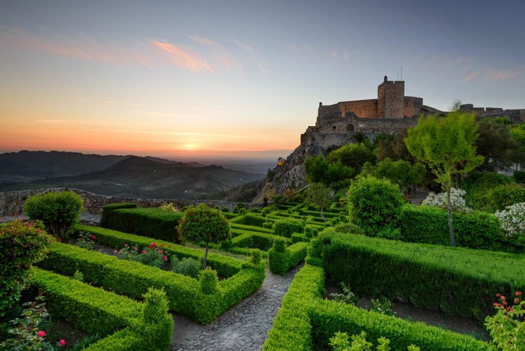 Scenic sunset view of a historic hilltop castle with lush green gardens and distant mountains in the background.