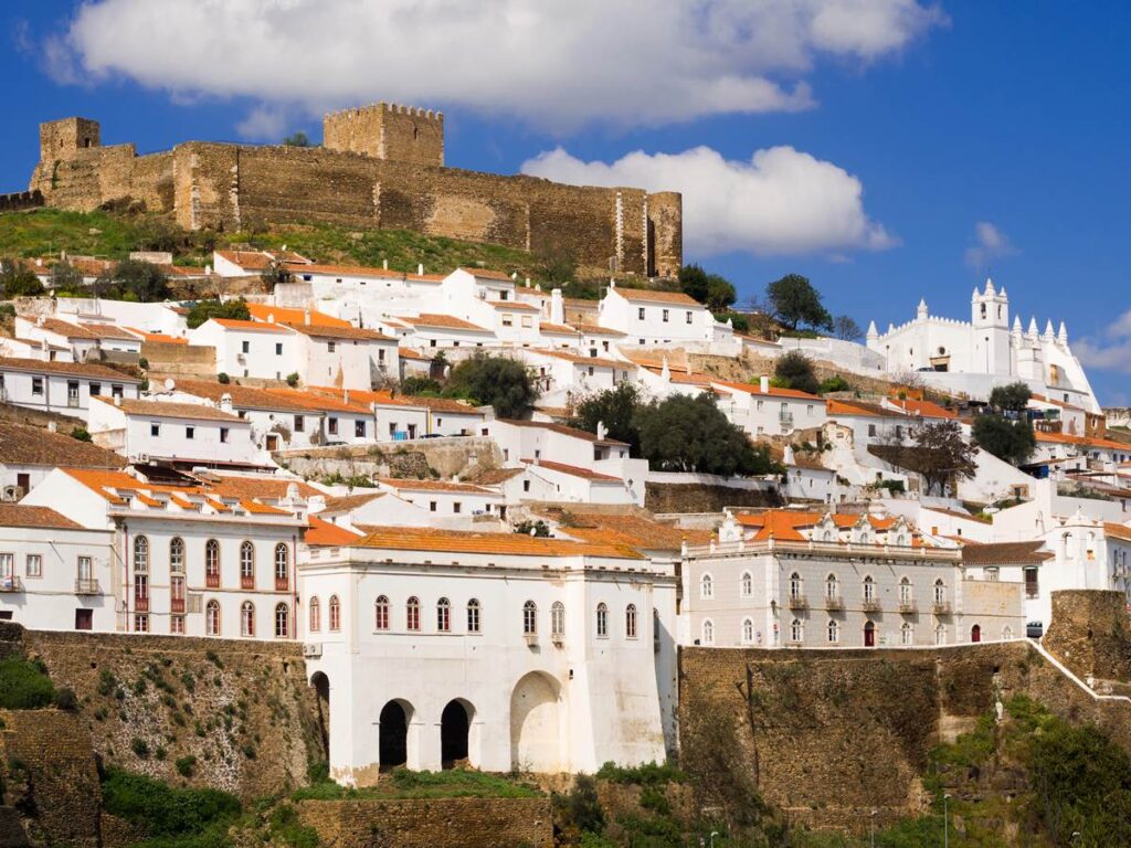 Scenic view of Mértola, Portugal, featuring historic castle and traditional whitewashed houses under a blue sky.