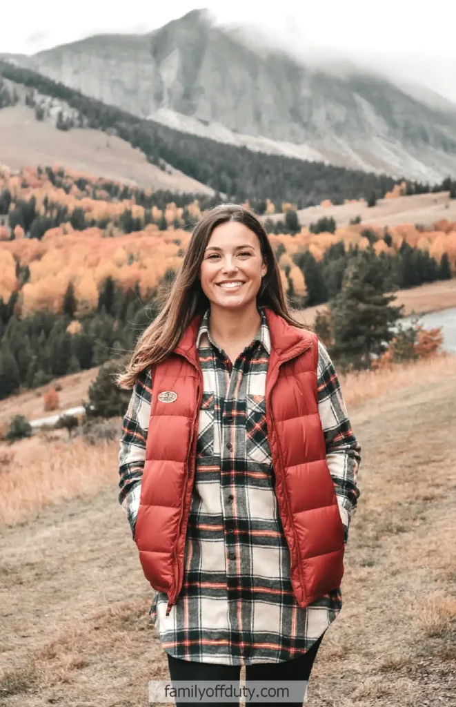 Smiling woman in plaid shirt and red vest, standing on a hill with colorful autumn foliage and mountains in the background.
