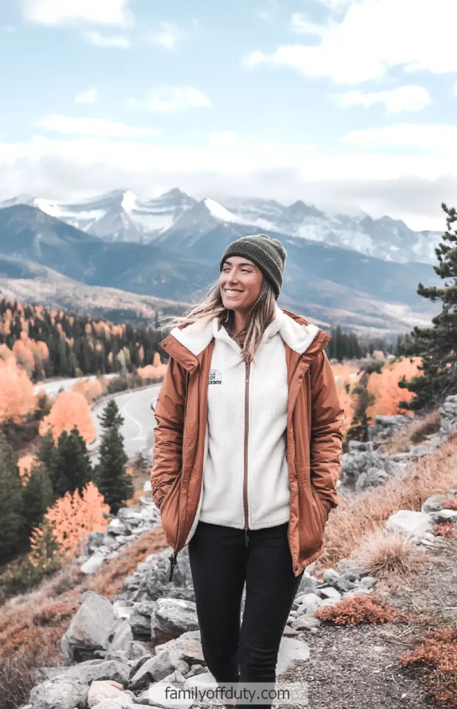 Smiling woman in a winter coat enjoys a scenic mountain view with fall foliage and snow-capped peaks.