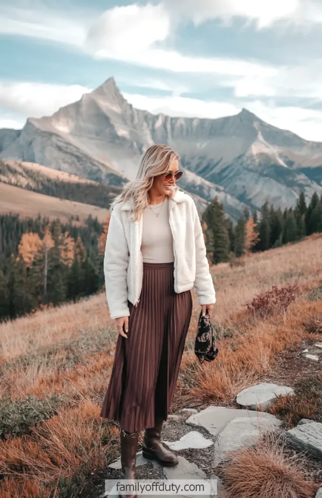 Fashionable woman in mountain landscape, wearing chic fall outfit with jacket, skirt, and boots.