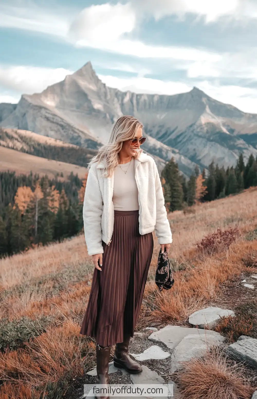 Mountain fall outfit ideas. Fashionable woman in mountain landscape, wearing chic fall outfit with jacket, skirt, and boots.