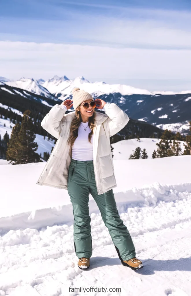 Smiling woman enjoying a snowy mountain view, dressed in winter gear with sunglasses and a beanie.