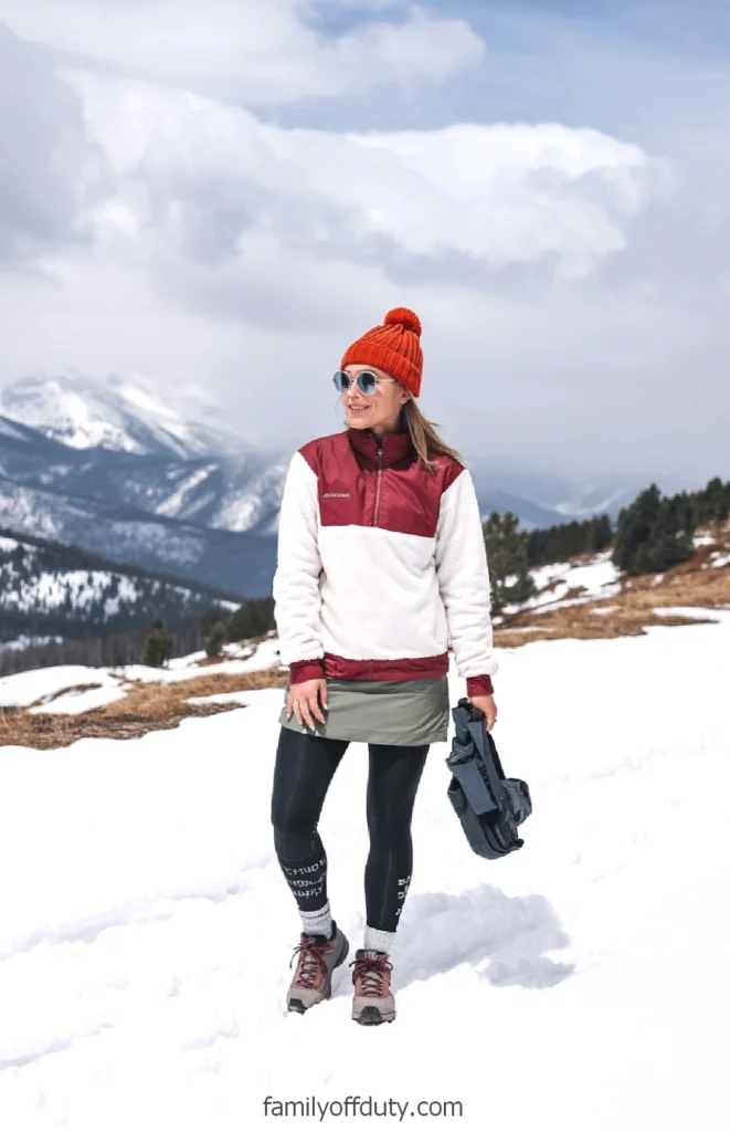Woman hiking in snowy mountain landscape, wearing winter attire and a red beanie, with scenic view in the background.