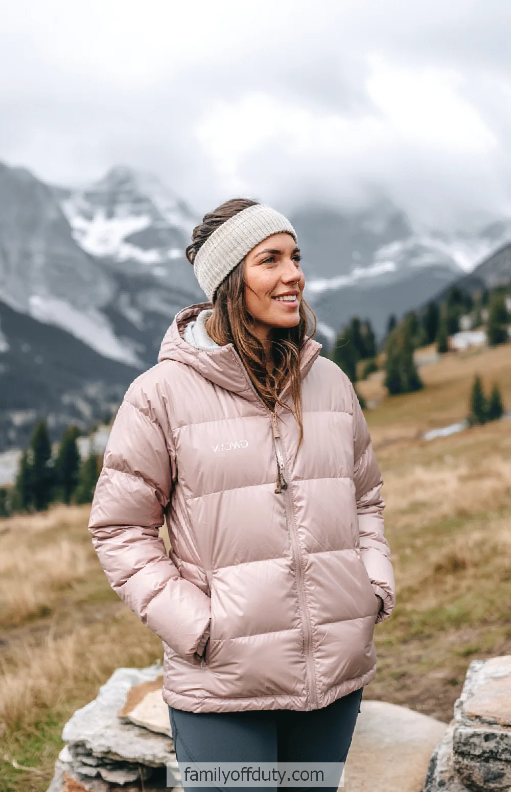 Mountain outfit ideas. Woman in pink puffer jacket smiling in scenic mountain landscape with snow-capped peaks and cloudy sky.