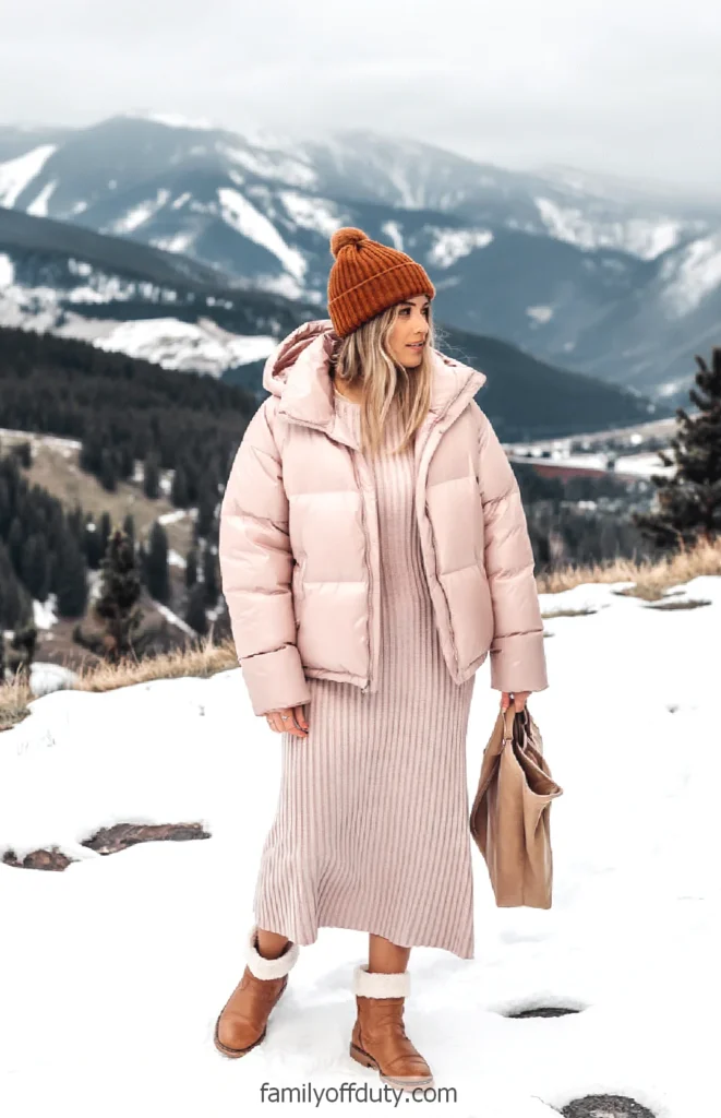 Woman in pink winter outfit and brown beanie standing on snowy mountain landscape, holding a beige tote bag.