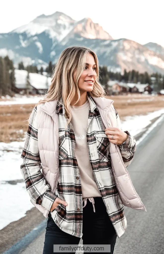 Woman in plaid jacket and vest enjoys a scenic winter backdrop of mountains and cabins.
