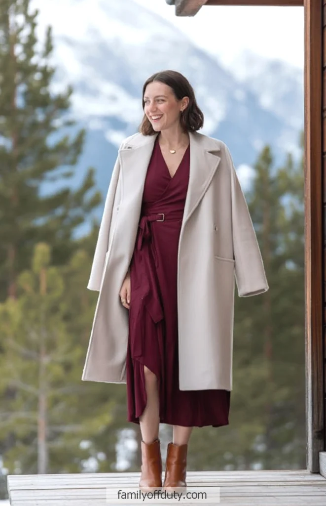 Woman in a burgundy dress and beige coat standing in a scenic snowy mountain setting.