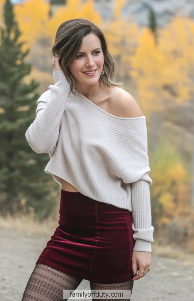 Woman in autumn fashion: off-shoulder sweater, burgundy skirt, smiling outdoors with fall foliage background.