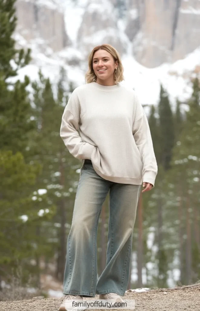 Woman in cozy sweater and jeans enjoying nature with snowy mountains in the background.