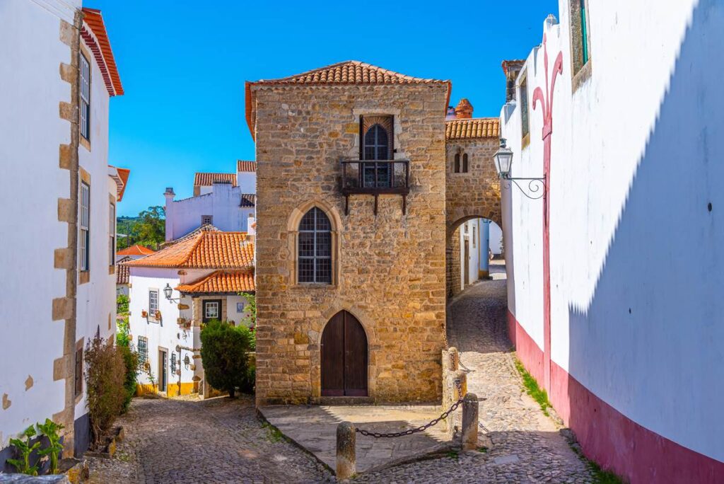 Medieval stone building with arched windows and a narrow cobblestone street in a historic village, under clear blue sky.