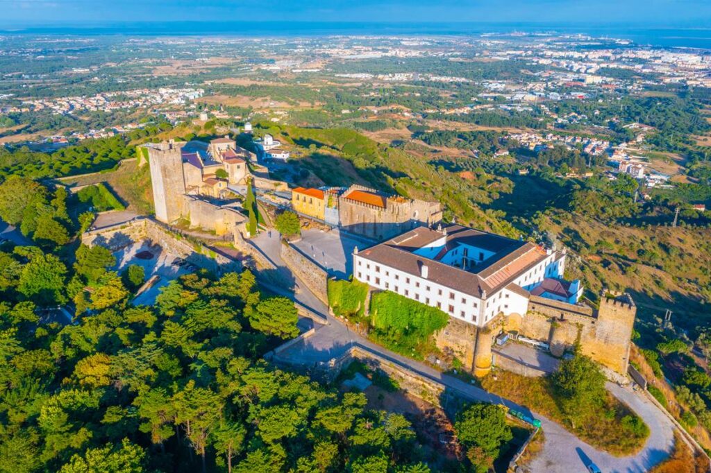 Aerial view of a historic castle surrounded by lush greenery and distant cityscape under a clear blue sky.