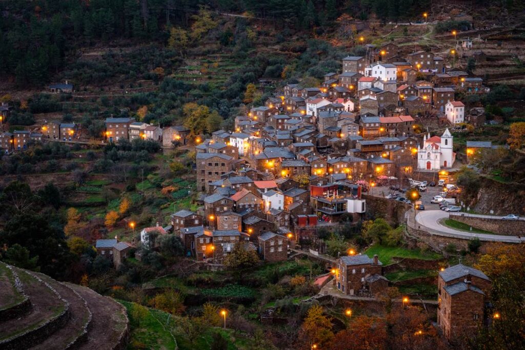 Charming hillside village at dusk with warm lights illuminating traditional stone houses and lush greenery.
