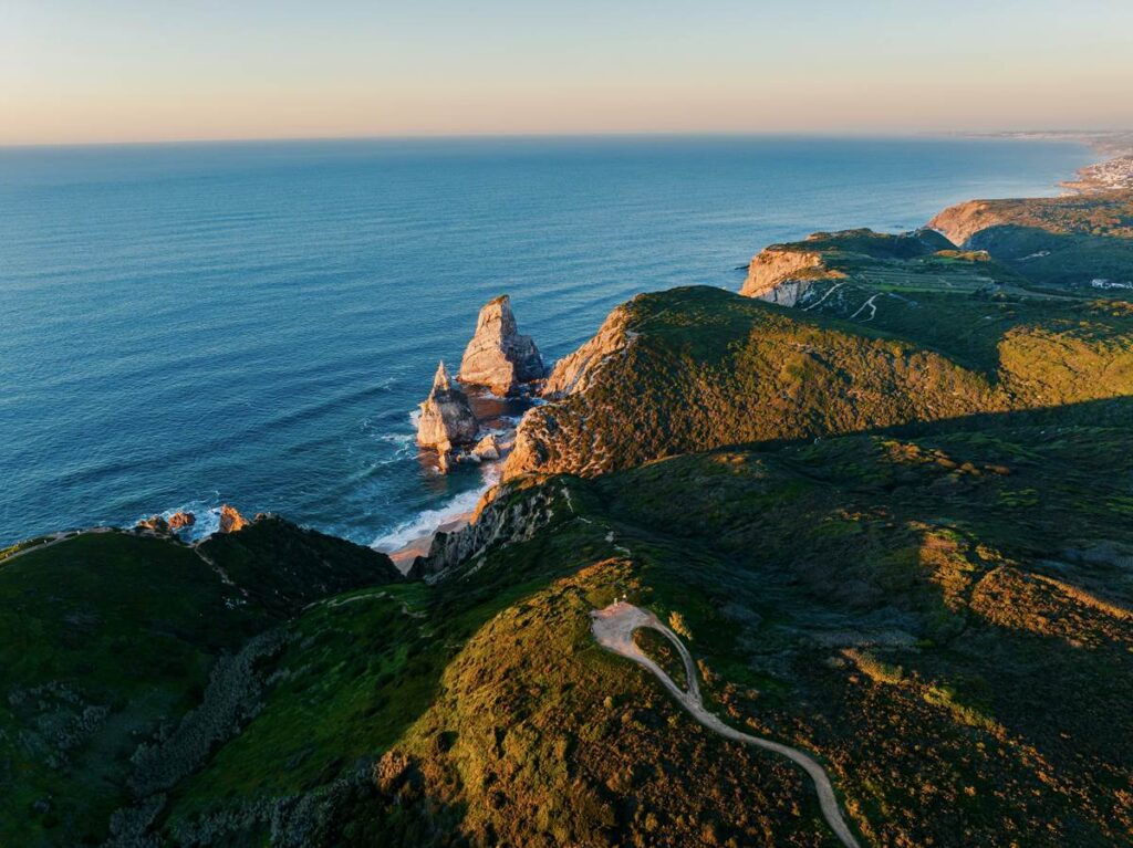 Aerial view of scenic coastline with cliffs and ocean waves at sunset, lush green landscape in foreground.