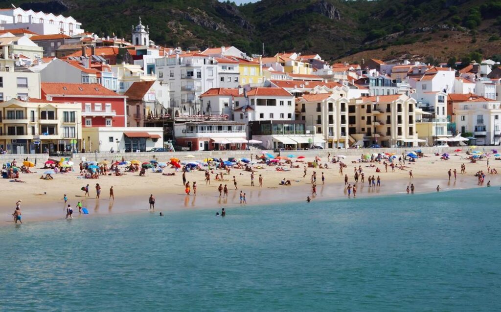 Sunny beach scene with colorful umbrellas, people enjoying the sand and sea, and charming town buildings in the background.