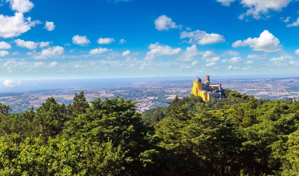 Colorful palace on a lush, green hilltop with a vast landscape view under a blue sky with clouds.