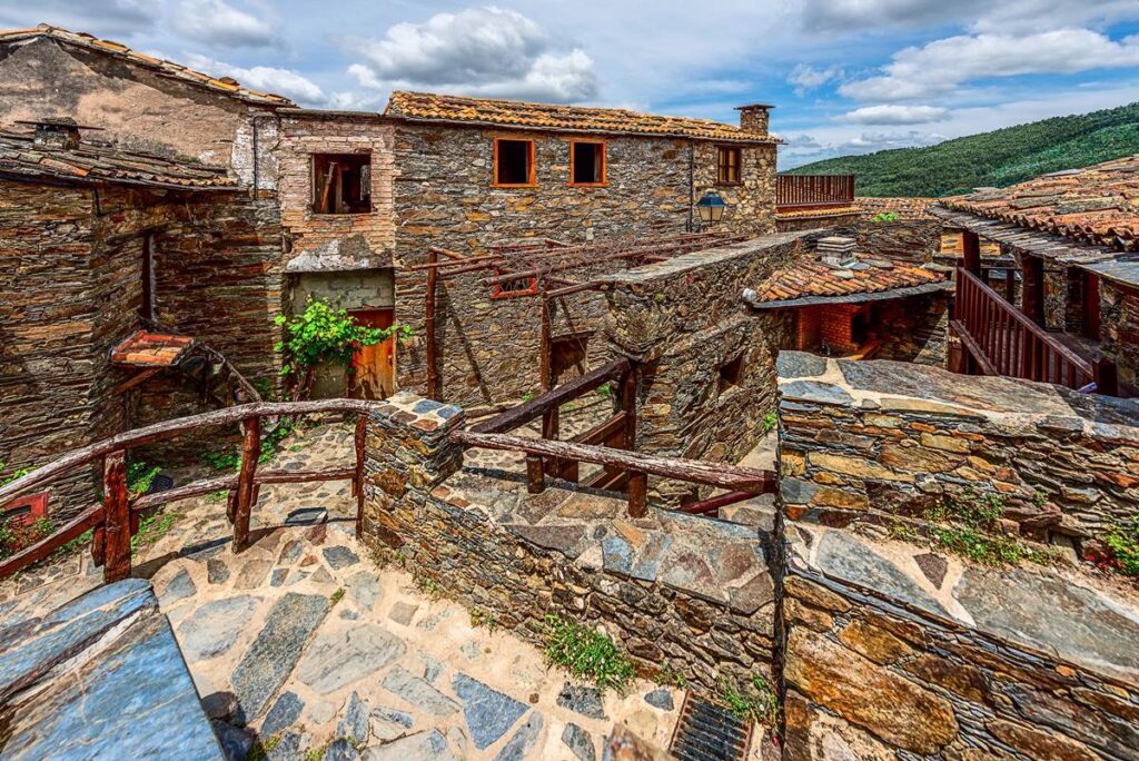 Historic stone village with rustic architecture under a blue sky, surrounded by green hillside.