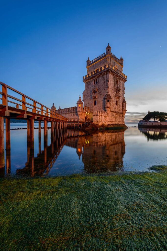 Historic Belém Tower in Lisbon, Portugal, reflected in water at sunset, with a clear blue sky above.