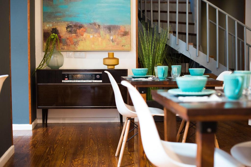 Modern dining area with blue tableware, abstract art, vintage radio console, plants, and wooden flooring under a staircase.