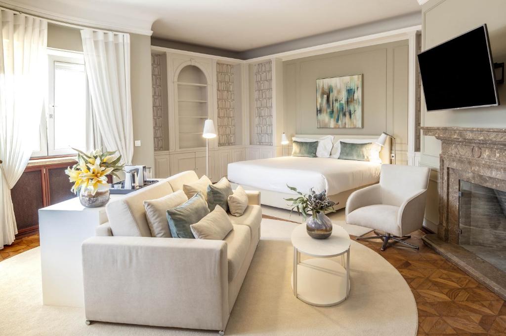 Elegant hotel room with cozy bed, modern decor, seating area, large window, and flat-screen TV near a fireplace.