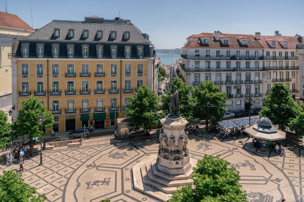 European square with a statue and mosaic pavement, surrounded by colorful buildings, trees, and a city view in the distance.