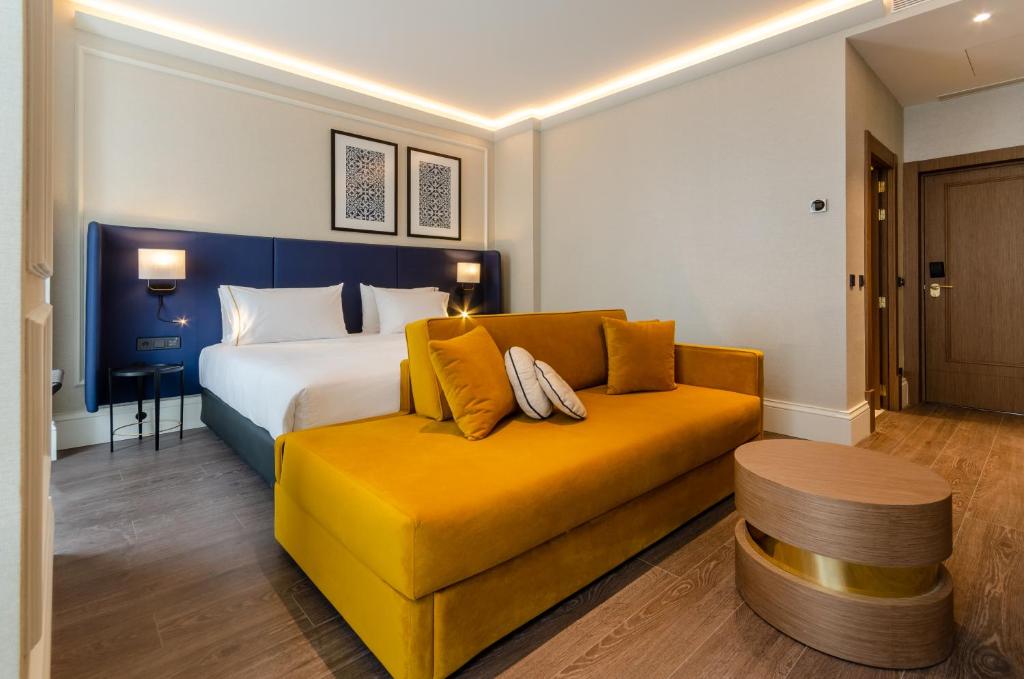 Modern hotel room with cozy double bed, vibrant mustard sofa, and stylish decor. Ideal for a relaxing stay.